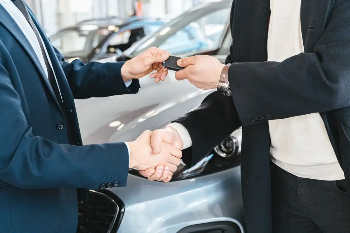 Pirkėjas ir pardavėjas spaudžia rankas prie automobilio ir perduoda raktelius.